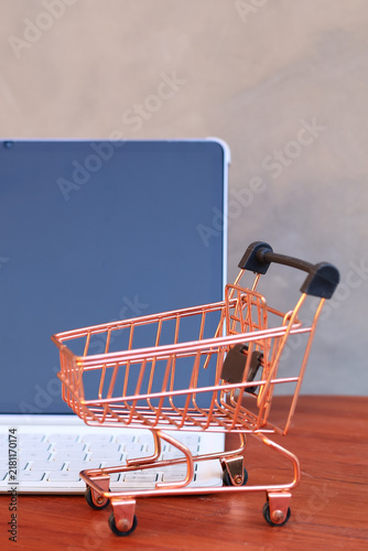 Miniature shopping cart in front of an open tablet with blank mockup screen on wooden table, online shopping and digital commerce concept.