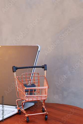 Miniature shopping cart in front of an open tablet with blank mockup screen on wooden table, online shopping and digital commerce concept.