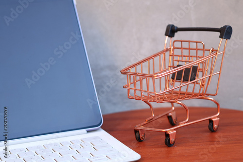 Miniature shopping cart next to a modern tablet with blank mockup screen on a wooden table, online shopping and e-commerce business concept with copy space.