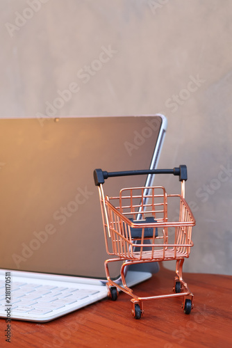 Miniature shopping cart in front of an open tablet with blank mockup screen on wooden table, online shopping and digital commerce concept.