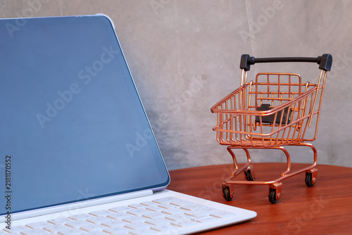 Miniature shopping cart next to a modern tablet with blank mockup screen on a wooden table, online shopping and e-commerce business concept with copy space.