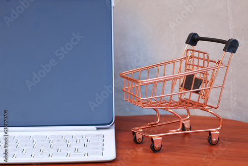 Miniature shopping cart next to a modern tablet with blank mockup screen on a wooden table, online shopping and e-commerce business concept with copy space.
