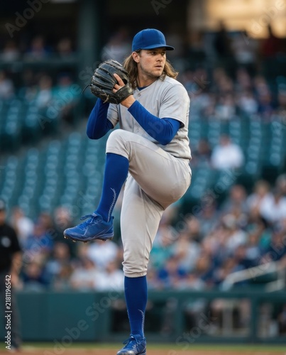 Professional baseball pitcher in a blue and grey uniform throwing a pitch on the mound during a game. Blurred stadium crowd in the background. High-energy sports action concept for commercial use.