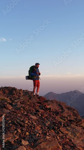 Alpinista con mochila de trekking ascendiendo por la cima de una montaña al atardecer con el sol detrás, luz dorada intensa y una vista panorámica épica del horizonte