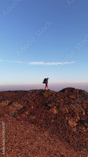 Alpinista con mochila de trekking ascendiendo por la cima de una montaña al atardecer con el sol detrás, luz dorada intensa y una vista panorámica épica del horizonte