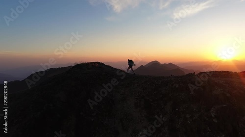 Alpinista con mochila de trekking ascendiendo por la cima de una montaña al atardecer con el sol detrás, luz dorada intensa y una vista panorámica épica del horizonte