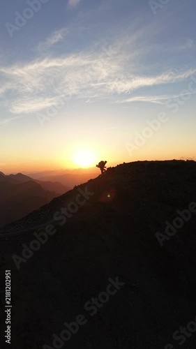 Alpinista con mochila de trekking ascendiendo por la cima de una montaña al atardecer con el sol detrás, luz dorada intensa y una vista panorámica épica del horizonte