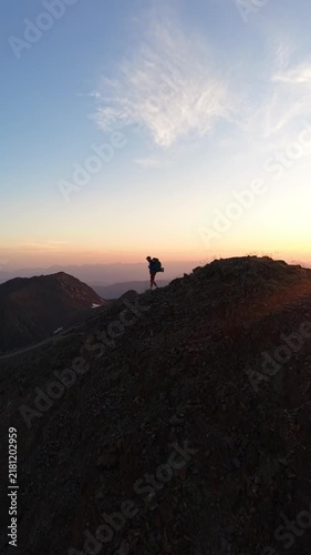 Alpinista con mochila de trekking ascendiendo por la cima de una montaña al atardecer con el sol detrás, luz dorada intensa y una vista panorámica épica del horizonte