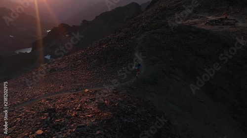 Alpinista con mochila de trekking ascendiendo por la cima de una montaña al atardecer con el sol detrás, luz dorada intensa y una vista panorámica épica del horizonte