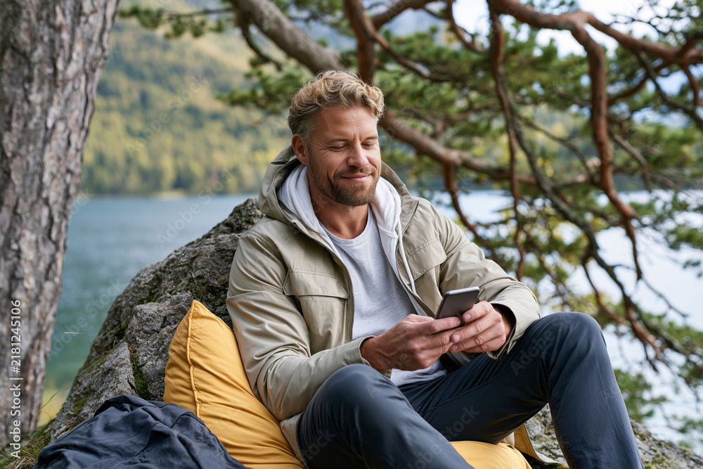Obraz premium Man relaxes on a yellow cushion beside a tree. He smiles while looking at his smartphone. Nature surrounds him with calm water and green hills. His coat and jeans suggest outdoor comfort