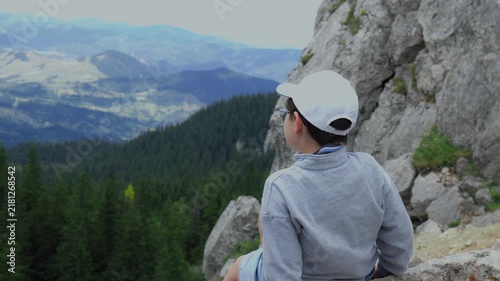 Young boy hiker in a white cap climbing rocks and enjoying a beautiful panoramic mountain view. Active childhood, summer trekking adventure, and outdoor nature exploration concept.
