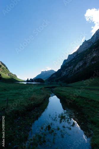 A small clear stream flowing through a shadowed green meadow towards Seealpsee lake surrounded by steep mountain peaks in the Appenzell Alps, Switzerland