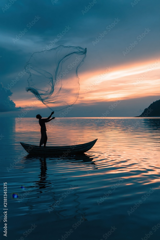 Obraz premium A fisherman in silhouette is captured mid action, casting a large circular net from a small boat on a still lake during sunset.