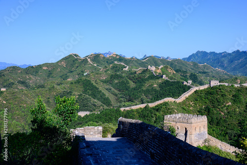 Foto The Jinshanling Great Wall, an ancient stone fortification, winds across lush green mountain slopes under a vivid blue sky, with steep stone steps and aged watchtowers rising amid dense vegetation