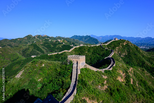 Canvastavla The Jinshanling Great Wall, an ancient stone fortification, winds across lush green mountain slopes under a vivid blue sky, with steep stone steps and aged watchtowers rising amid dense vegetation