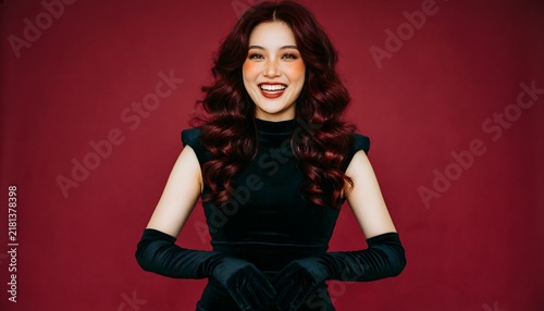 A beautiful young woman with curly hair wearing a black dress and elegant gloves posing against red background