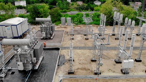 Aerial drone view of an electrical substation under construction in Sumy, Ukraine. High-voltage transformers, switchgear, transmission equipment, utility buildings, workers, and a yellow excavator