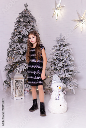Elegant Little Caucasian Girl in Sequin Dress Posing Near Snowy Christmas Trees with Snowman and Star Decor