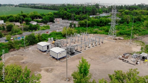 Aerial drone view of an electrical substation under construction in Sumy, Ukraine. High-voltage transformers, switchgear, transmission equipment, utility buildings, workers, and a yellow excavator