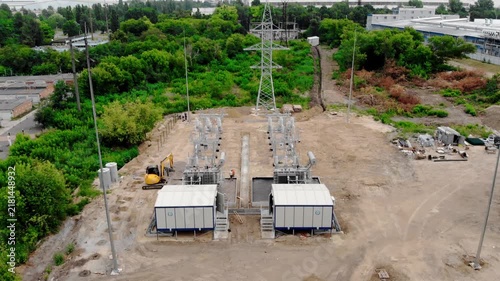 Aerial drone view of an electrical substation under construction in Sumy, Ukraine. High-voltage transformers, switchgear, transmission equipment, utility buildings, workers, and a yellow excavator