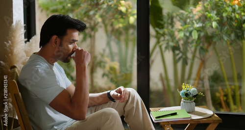 Indian calm young adult gen z relax man sitting on chair hold tea mug drink coffee enjoy day room male boy spend time take casual job break home house place
