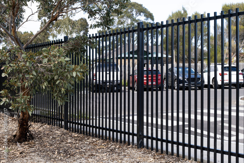 A black steel palisade security fence bordering a suburban car park in Australia, with parked vehicles visible through the fence rails. Commercial boundary or institutional perimeter fencing