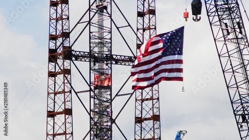 United states flag attached to a crane hook, majestically waving in the wind in slow motion with a large construction site lattice structure in the background