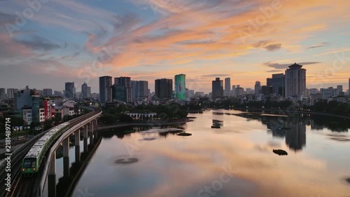 Elevated Cat Linh–Ha Dong metro train curves beside Ho Dong Da in Hanoi, Vietnam, with a calm lake, urban skyline, and colorful sunset reflections.