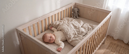 A cute baby sleeps peacefully under a soft blanket inside a wooden crib in a sunlit nursery room, calm morning routine, sweet infant nap, safe family home, gentle nursery moment.