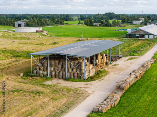 Aerial view of a farm featuring a large open-sided shed stacked with round hay bales, with additional bales lined up along a dirt road in the rural landscape of Virpe, Ventspils novads, Latvia.