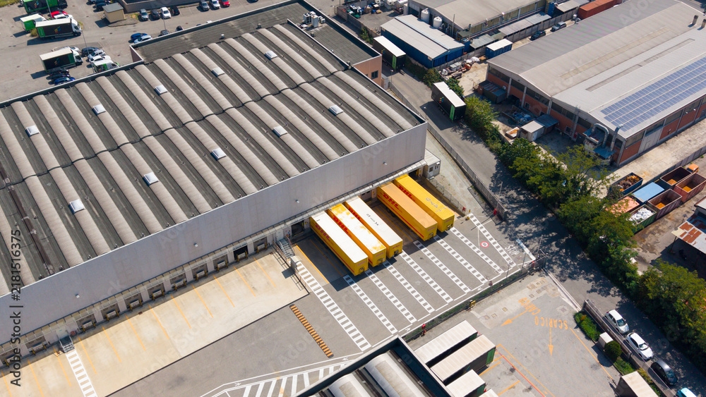 © Amazing Aerial - San Giuliano Milanese, Italy - 04 September 2026: Aerial view of the DHL Express facility with yellow delivery trucks parked at the loading bays of a large industrial warehouse.