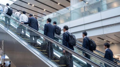 Businessmen ascending escalator in modern station, busy commuters moving upward in formal attire with briefcases and backpacks