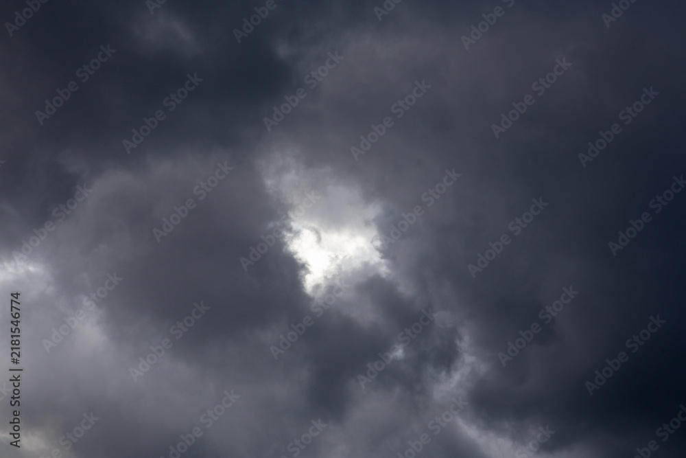 Fototapeta premium Dramatic dark storm clouds filling the sky with a small bright white sun light break. Overcast weather scene before a heavy rain. Natural atmospheric backdrop and texture template for graphic design. 