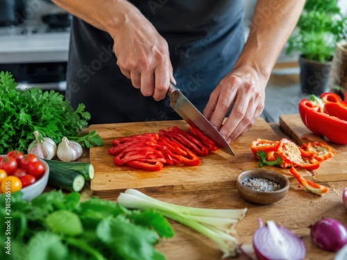 Chef prepares fresh vegetables on wooden cutting board in a kitchen setting with green herbs and other ingredients nearby