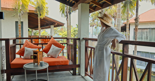 фотографія Woman in white bathrobe and straw hat standing on wooden balcony, tropical resort terrace, luxury vacation lifestyle, travel leisure, commercial hospitality industry