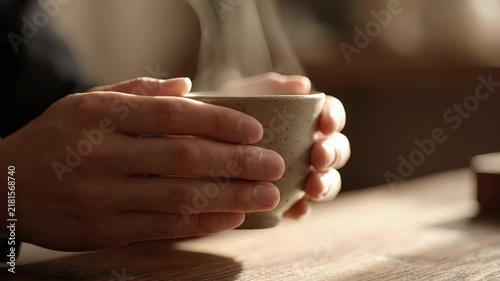 Hands holding a steaming hot cup of tea or coffee on a wooden table in a cozy cafe setting.