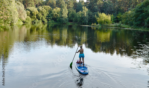 Картината върху платно Individual paddleboarding on calm lake surrounded by lush greenery and trees, re