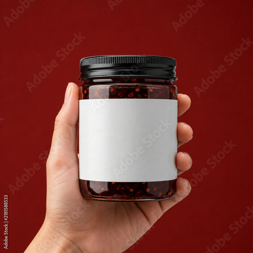 Clear glass jar mockup with blank white label held by human hand against solid dark red background, concept for condiment branding design and product packaging showcase, ideal for chili sauce