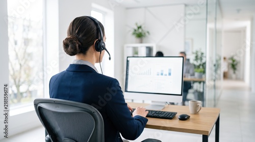 Female customer support agent wearing headset working at computer in modern bright office, professional call center operator providing assistance, business communication service