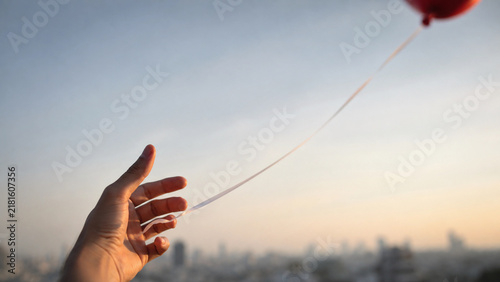 Hand gently releasing red balloon string against soft sunset sky with blurred city skyline, symbolizing letting go, moving forward, freedom, and emotional release