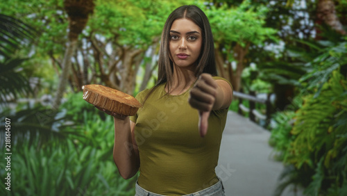 Cuadro en lienzo Young hispanic woman holding a cake base and showing thumbs down on a forest walkway with direct gaze and neutral expression; disapproval critique