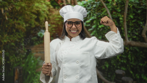 Foto Woman chef wearing toque holds rolling pin and flexes arm with fist, smiling toward camera in forest; confidence empowerment skill