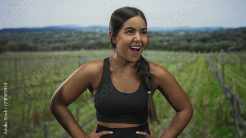 Sporty woman with hands on hips smiling widely in a vineyard, wearing black sports bra and braid visible; confident athletic energy.
