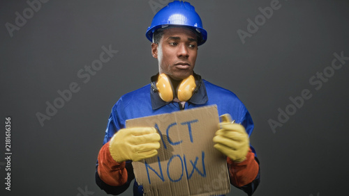 Obraz na płótnie Man wearing blue hardhat and work coveralls holds cardboard sign reading act now and gestures with gloved hand in a studio; labor rights urgent