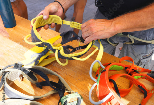 Hands adjusting yellow climbing harness on wooden table. Multi colored safety gear preparation for rock climbing outdoor activity. Instructor inspecting protection equipment for mountaineering sports.