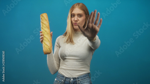 Obraz na plátně Young redhead woman holds baguette in left hand and shows palm out in stop gesture to camera in studio; rejection food refusal
