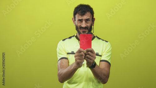 Fotografie Referee man with beard holds red card in hand, grimaces and raises other hand while wearing whistle and headset in studio; discipline frustration