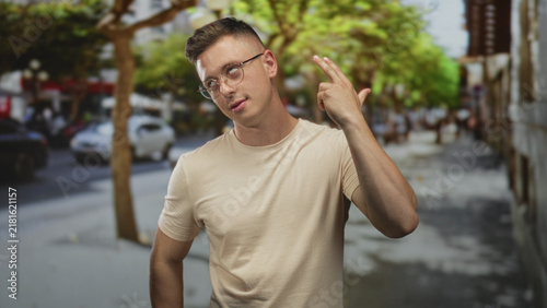 Man wearing glasses points finger, other hand on hip on a busy street with trees and parked cars; thoughtful calm.