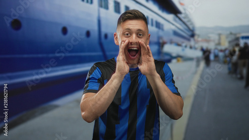 Foto Man cupping hands to mouth shouting on street beside a ship and pier, wearing blue black soccer jersey and smiling; excitement