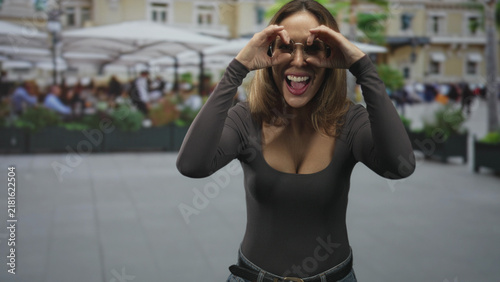Fotografía Woman making binocular hand gesture with hands and glasses, smiling with cheerful expression on a street cafe; playful joy